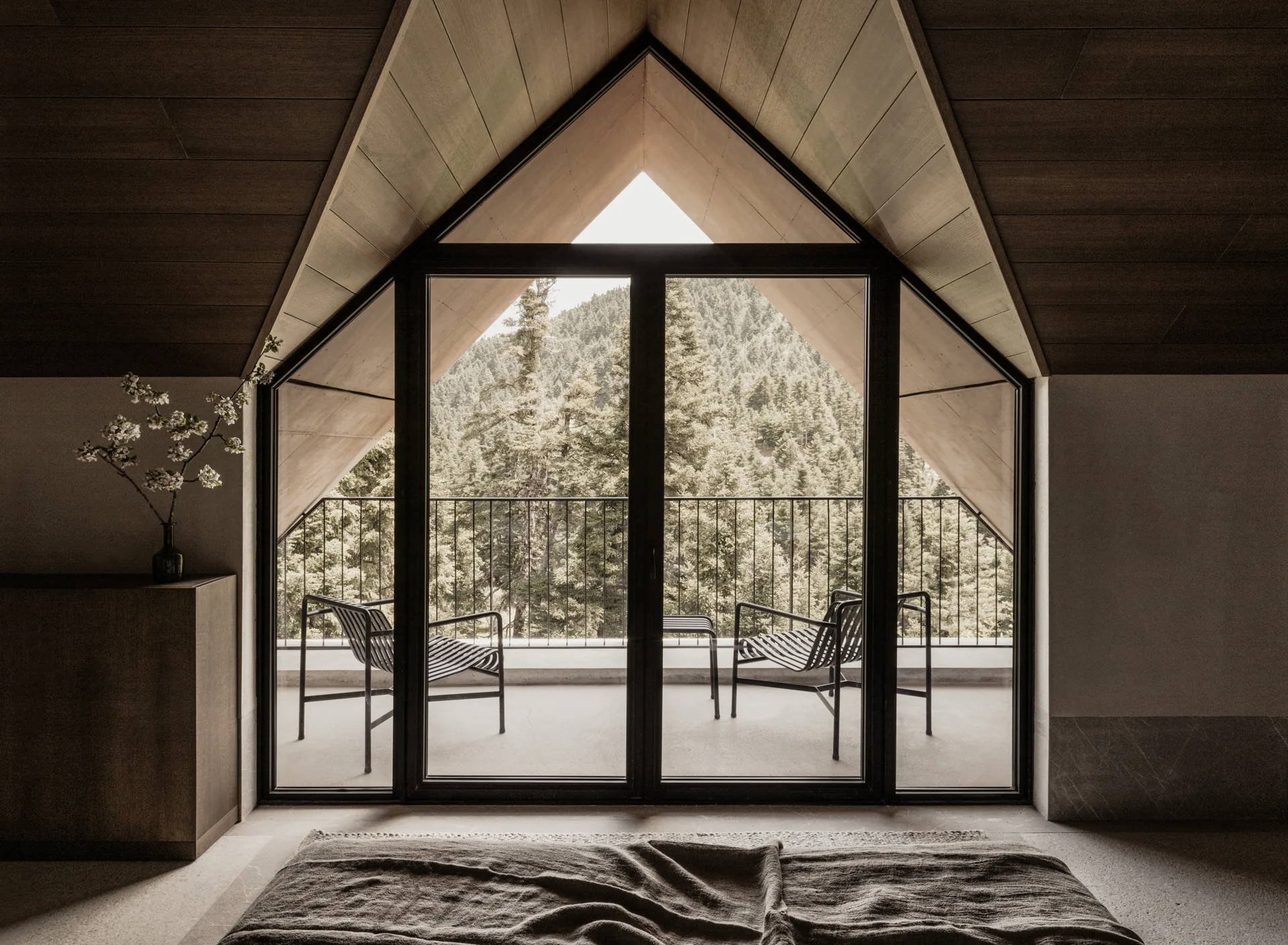 A bed beneath a timber-lined gable, with a balcony and fir-covered mountain beyond the glass