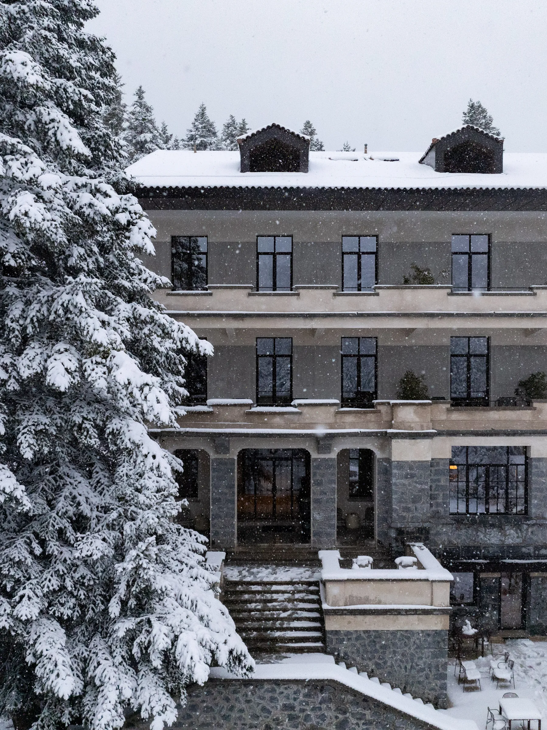 MANNA's stone entrance and terrace during snowfall, with a snow-covered fir beside the steps