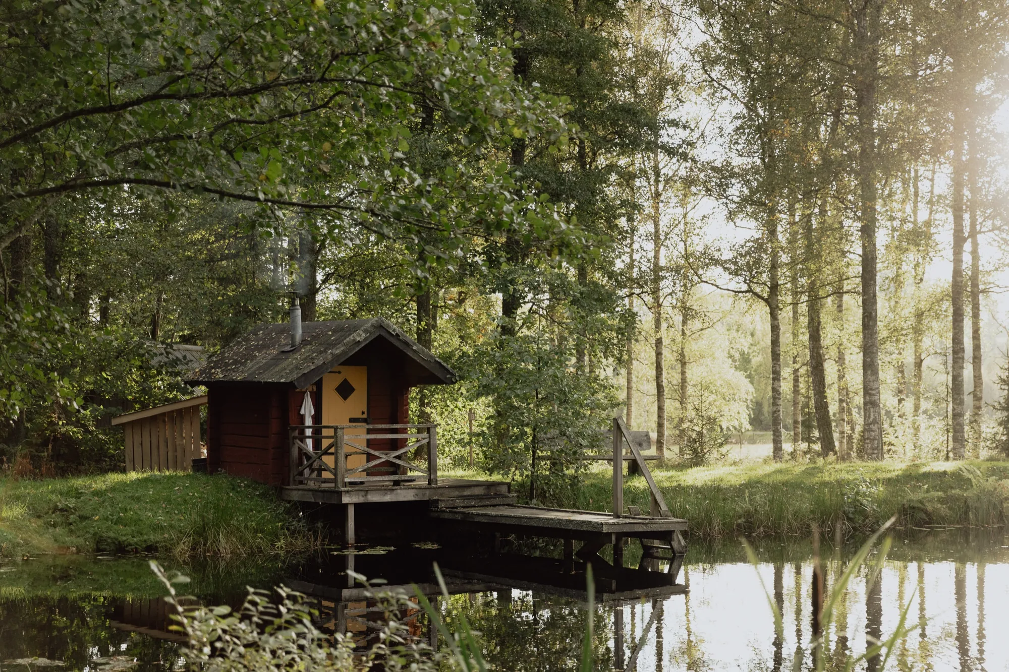 Small red timber sauna with a yellow door on a jetty beside a calm forest lake