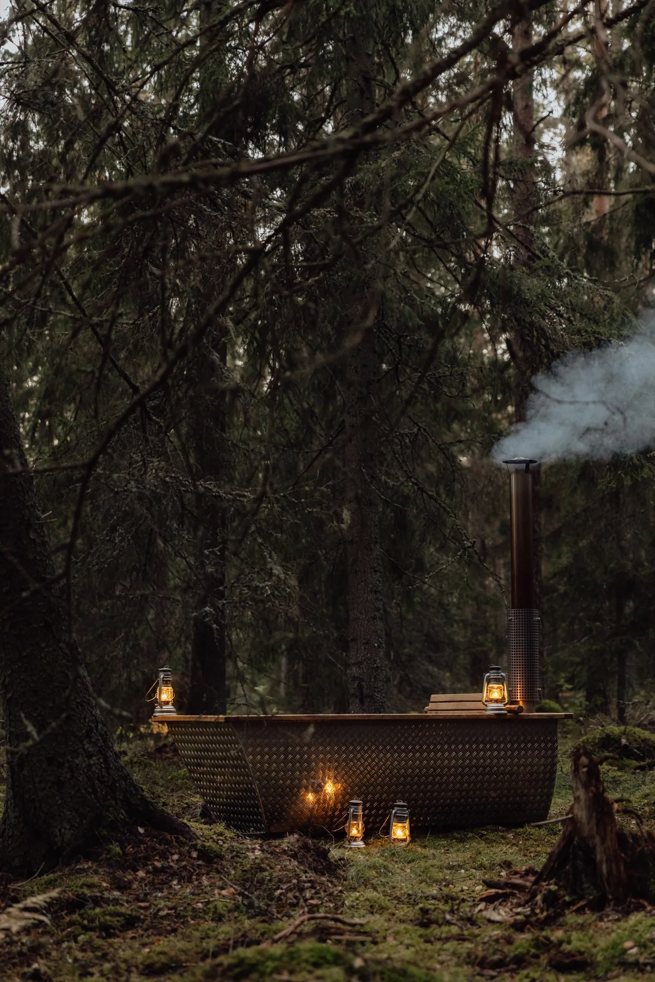 Outdoor metal bath with a timber rim, lanterns and rising smoke in the dark spruce forest