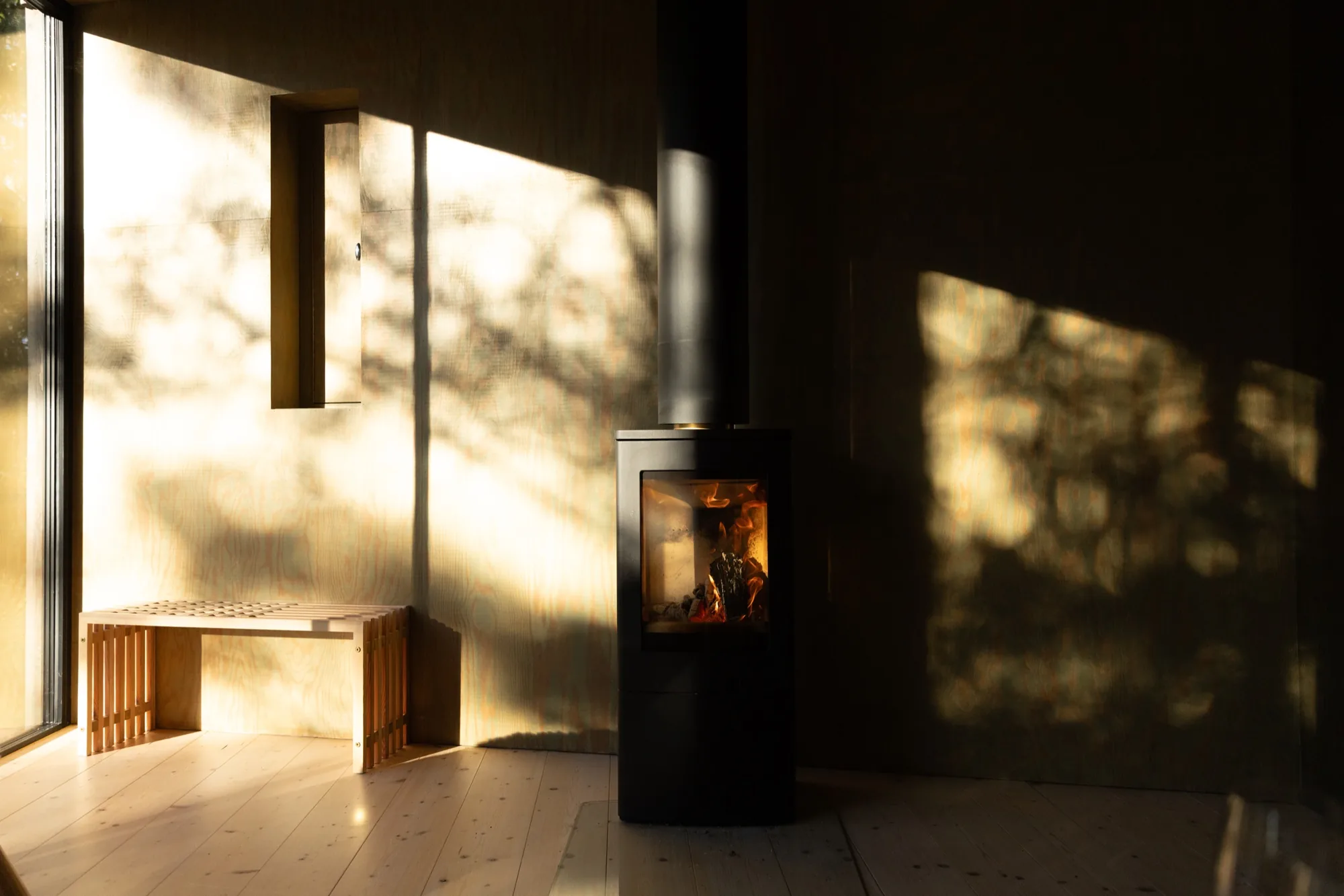 Black wood-burning stove beside a pale timber bench, with tree shadows crossing the interior
