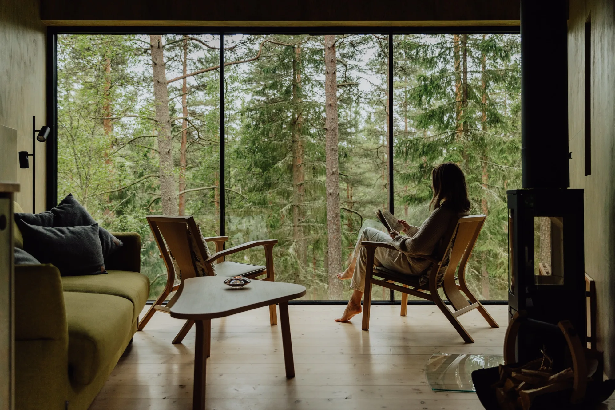 Woman reading barefoot in a Forest Suite living room with full-height windows onto the spruce woods