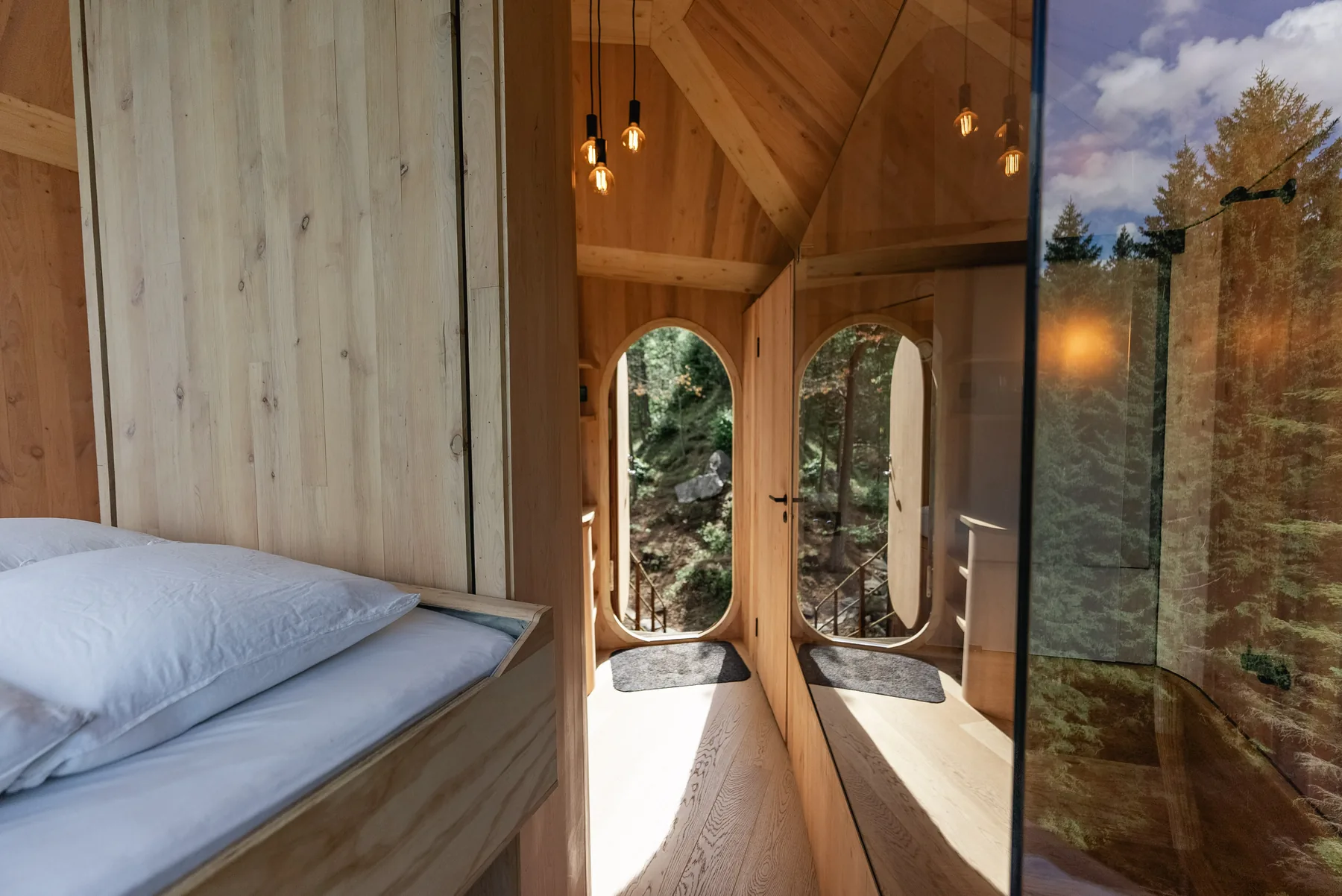 Warm timber sleeping area with oval doors and forest reflected in the glass-walled bathroom